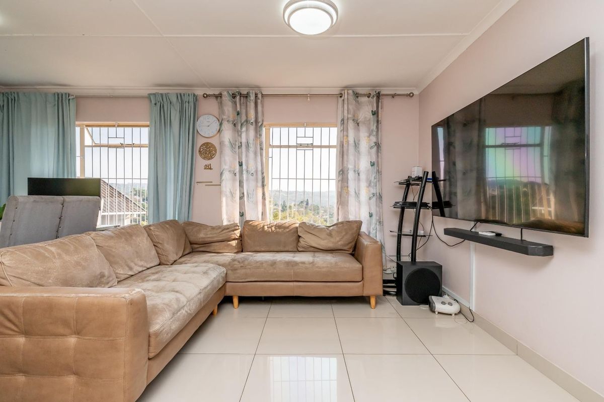 A well-lit living room featuring large windows with security bars, neutral tiled flooring, and a clean, functional layout.