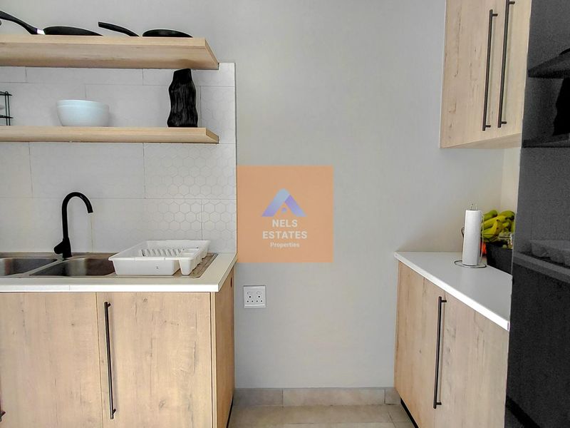 A modern kitchen featuring light wood-finish cabinetry, white countertops, a tiled backsplash, and open shelving with a functional sink area.