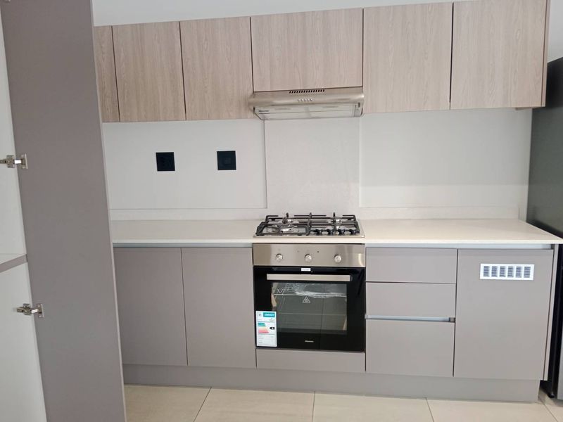 A modern, clean kitchen featuring light wood-tone upper cabinets, grey lower cabinetry, a gas hob, built-in oven, and a sleek stone countertop.