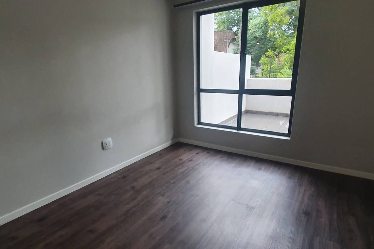 A bright, empty bedroom featuring dark laminate flooring, neutral walls, and a large window providing natural light and access to an outdoor balcony area.