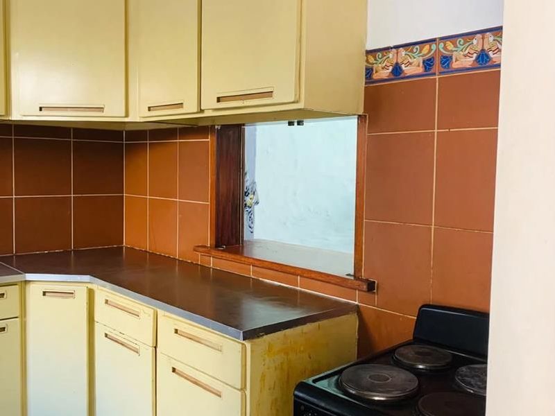 A dated kitchen featuring yellow cabinetry, brown tiled backsplash, and a convenient pass-through window to the adjacent living area.
