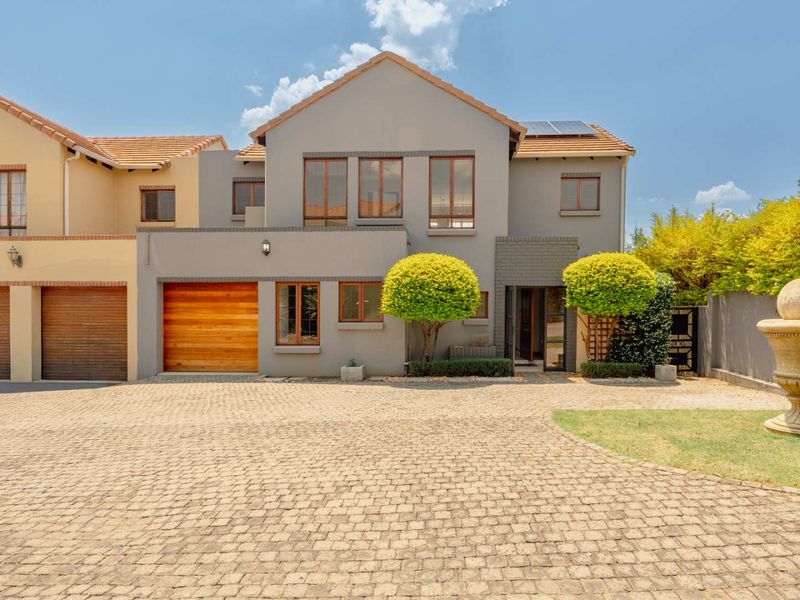 A well-maintained double-story cluster home featuring a paved driveway, wooden garage door, solar panels, and a decorative stone fountain in the front garden.