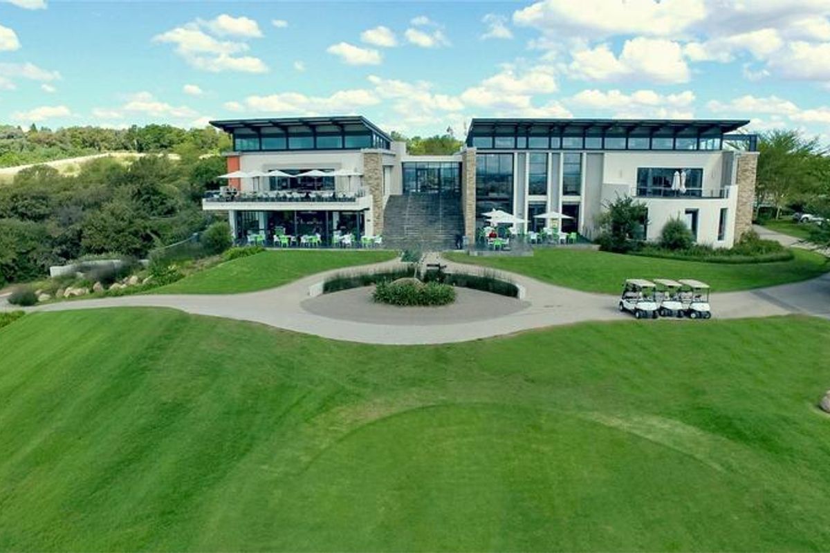 An aerial view of a modern clubhouse building featuring expansive glass facades, tiered outdoor terraces, and manicured green landscaping.