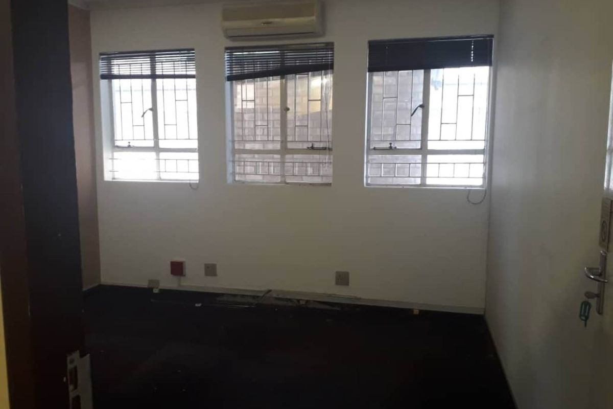 An empty room featuring three windows with security bars, a wall-mounted air conditioning unit, and dark flooring in need of repair.