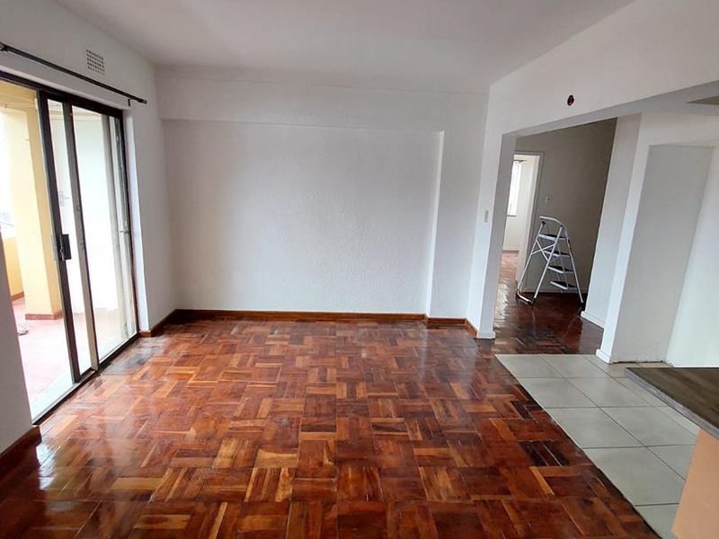 An empty open-plan living area featuring polished parquet flooring, a sliding glass door leading to a balcony, and neutral wall finishes.
