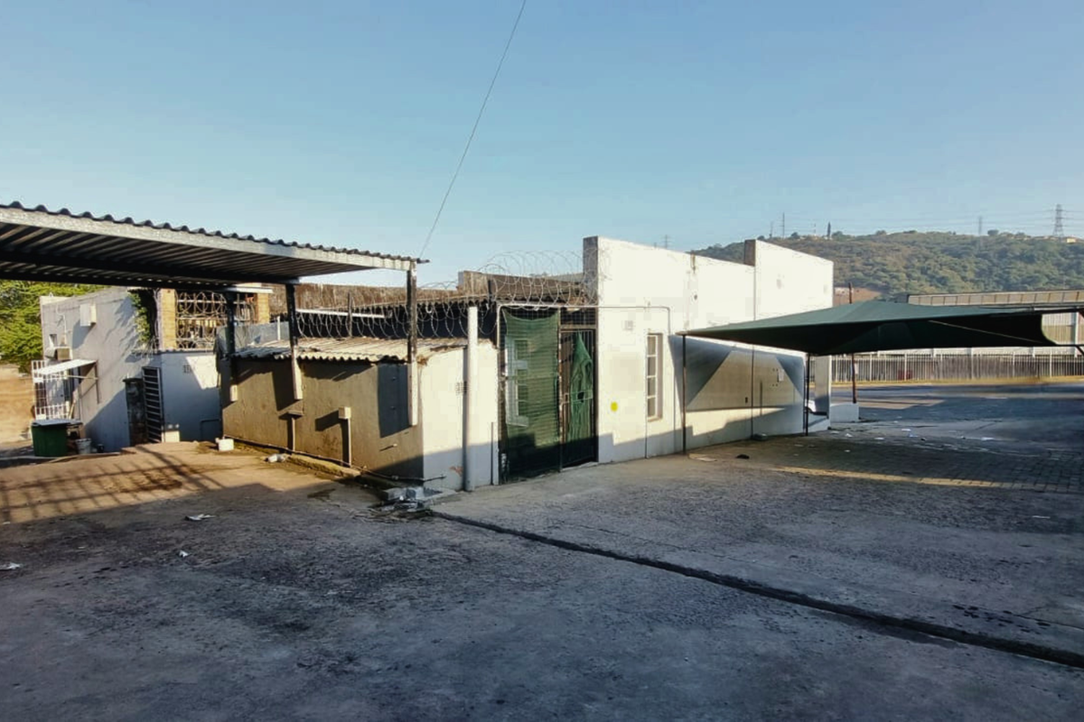 Rear exterior view of a commercial property featuring concrete yard space, metal carports, shade sails, and security fencing.