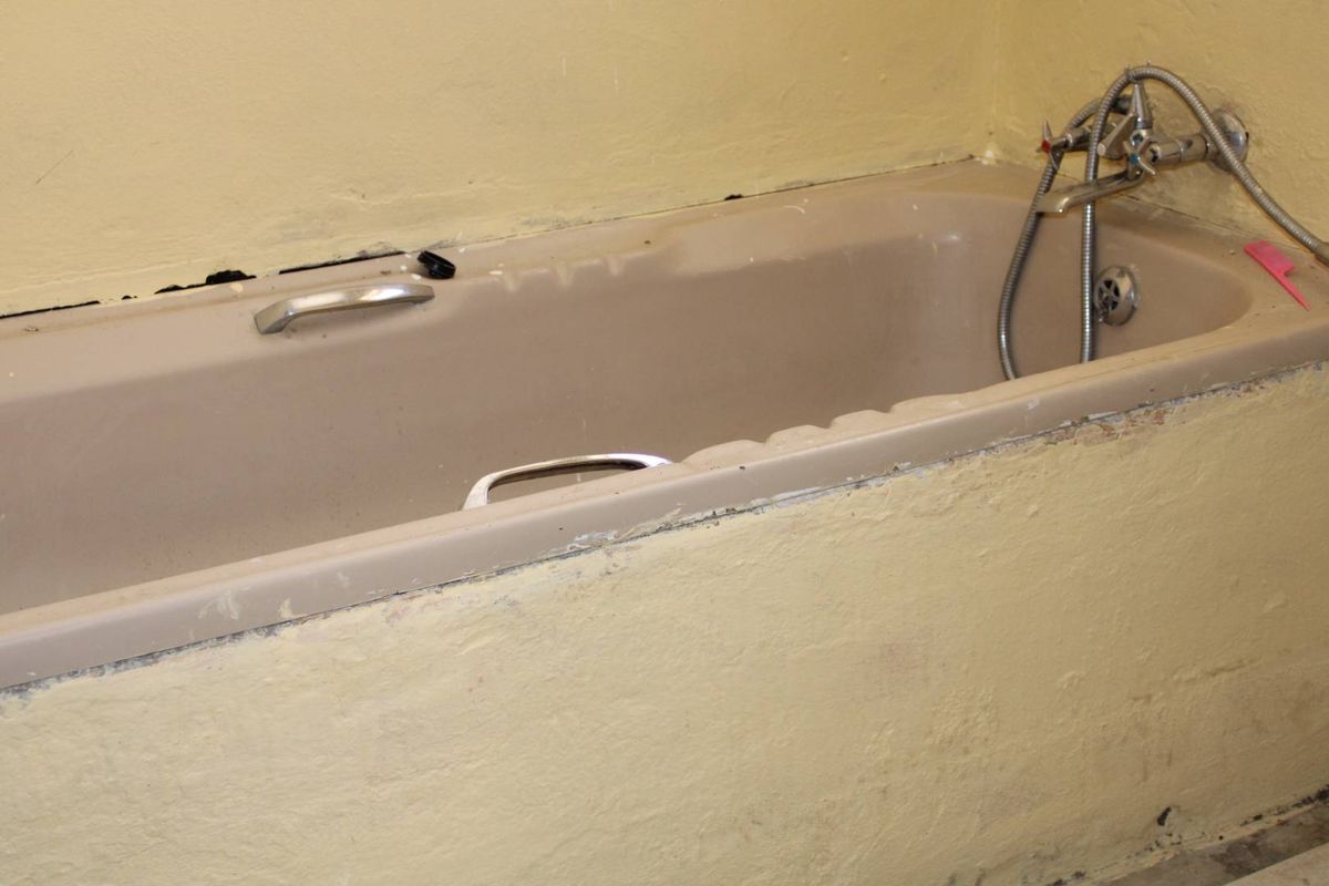 A dated bathroom featuring a built-in bathtub with handheld shower fixtures, requiring cosmetic updates and repairs to the surrounding wall finishes.