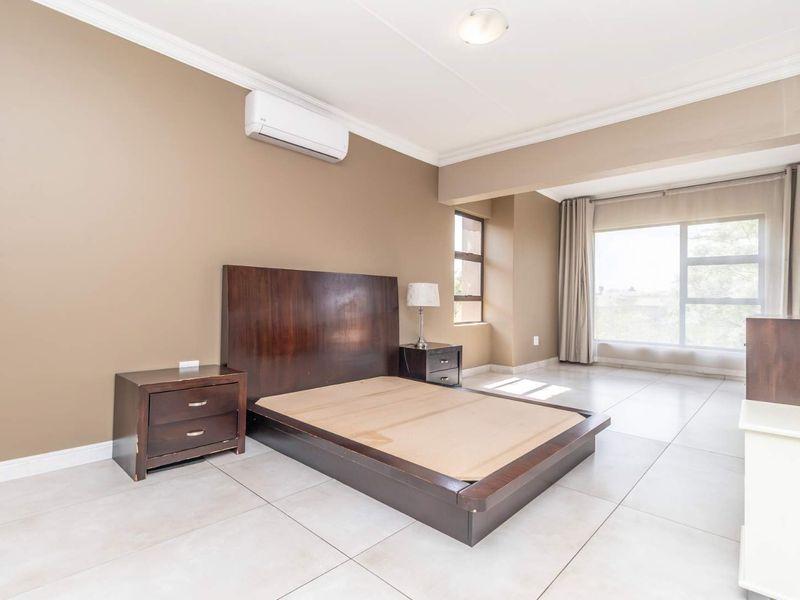 Spacious bedroom featuring neutral wall tones, large windows for natural light, tiled flooring, and a wall-mounted air conditioning unit.