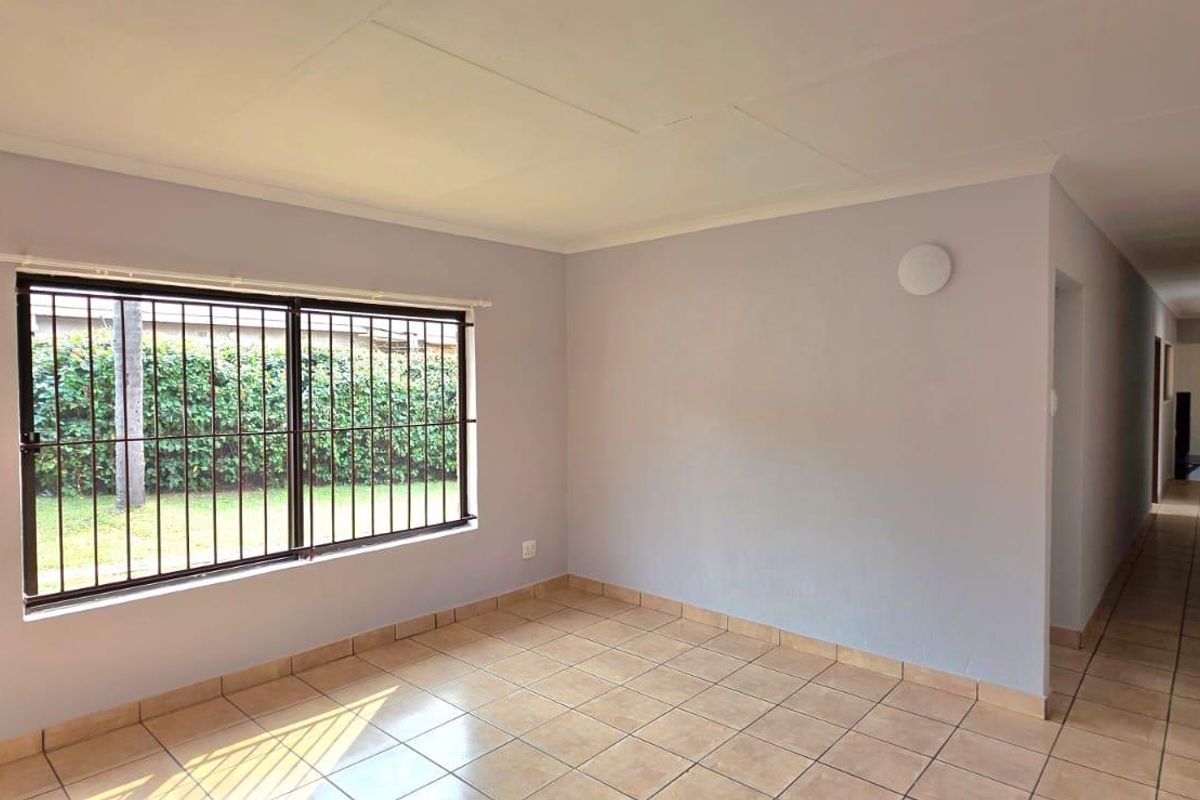 A bright, empty living room featuring neutral walls, tiled flooring, and a large window with security bars, leading into a long hallway.