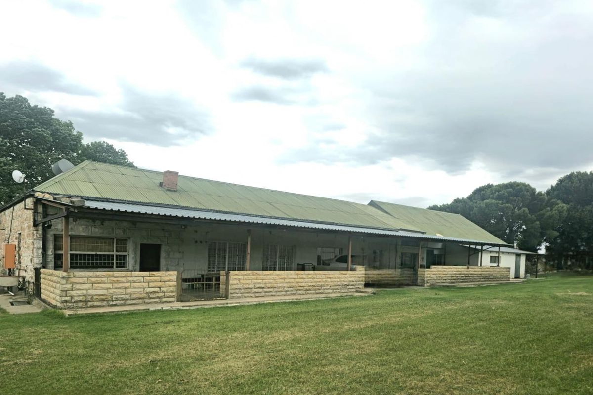 A rear view of a farm-style dwelling featuring a long covered patio, stone masonry walls, and a corrugated metal roof set against a large lawn.