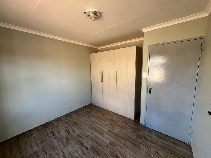 A clean, empty bedroom featuring modern wood-look floor tiles, neutral wall paint, and a built-in wardrobe with light wood finish.