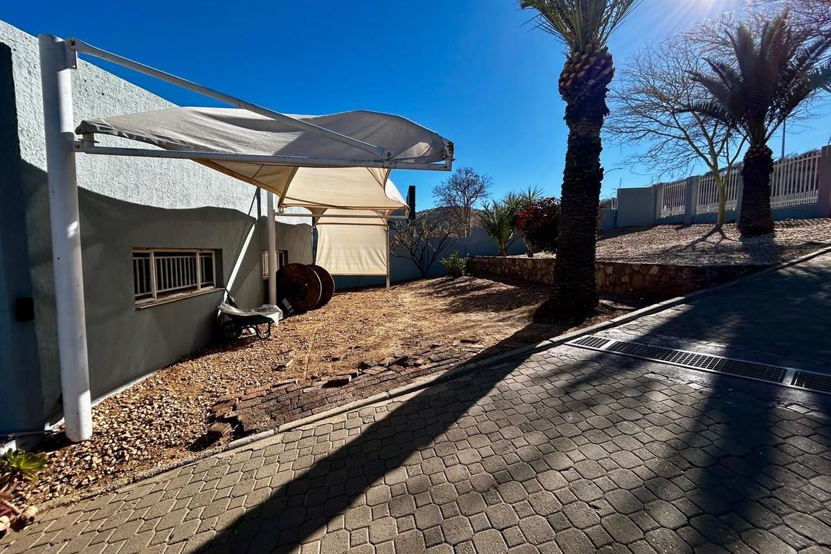 Rear exterior area featuring a paved driveway, shade sail structure, and a landscaped garden with a retaining wall.
