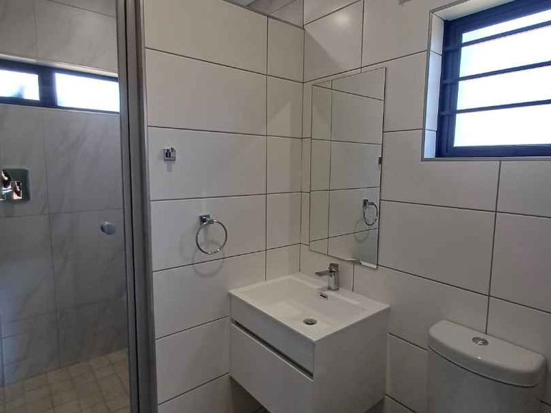 A modern, clean bathroom featuring a glass-enclosed shower, wall-mounted vanity with basin, and a standard toilet with neutral wall and floor tiling.