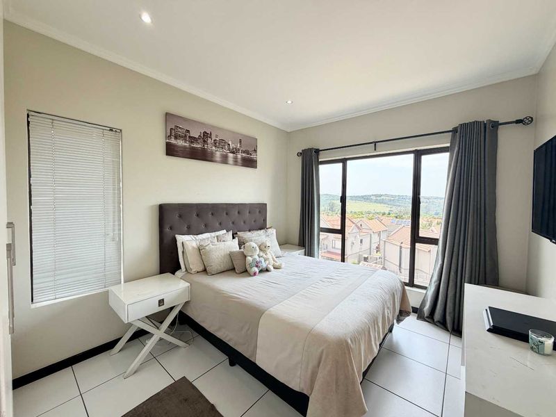 A well-maintained bedroom featuring tiled flooring, recessed lighting, and a large window with blinds and curtains, offering pleasant views of the surrounding neighborhood.