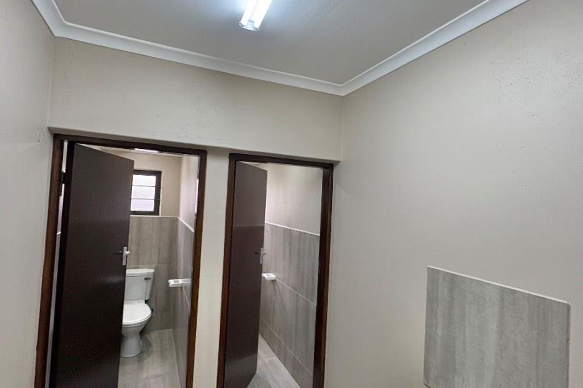 A clean, functional office bathroom featuring a wall-mounted basin and separate toilet cubicles with neutral tiled flooring and modern finishes.
