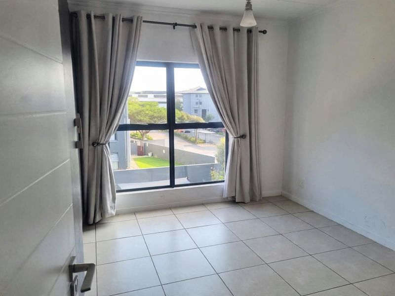 A bright, empty bedroom featuring neutral tiled flooring, a large window with curtain fixtures, and a clean, modern interior finish.