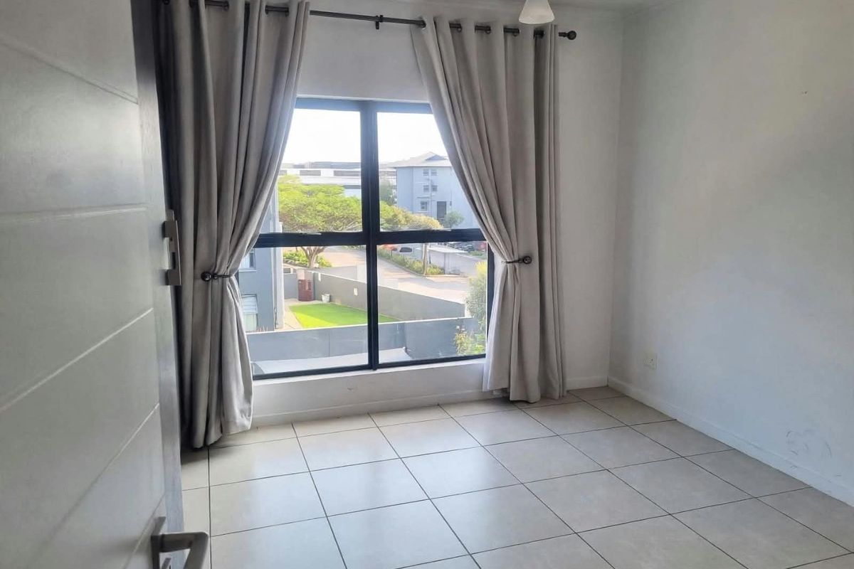 A bright, empty bedroom featuring neutral tiled flooring, a large window with curtain fixtures, and a clean, modern interior finish.