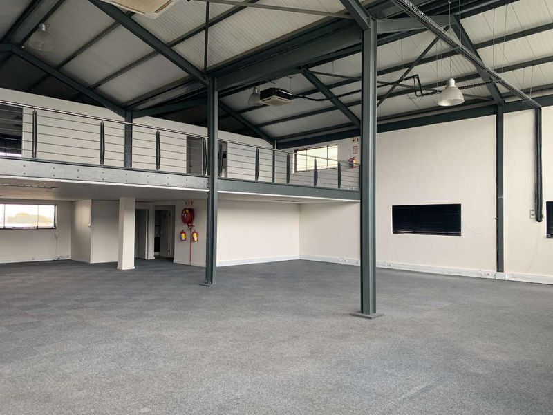 Spacious open-plan office area featuring a mezzanine level, exposed steel structural elements, and neutral carpeted flooring in a well-maintained commercial setting.