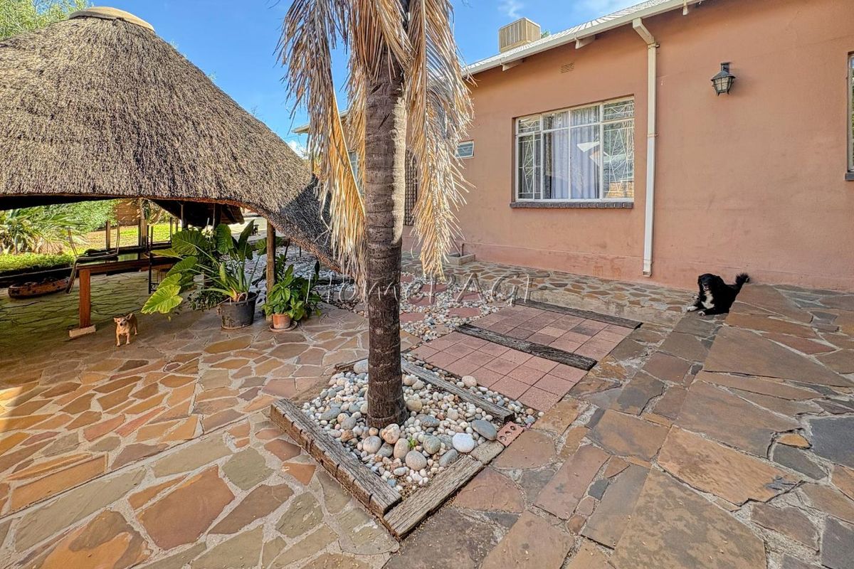 A rear exterior view featuring a paved patio area, a thatched lapa structure, and established garden landscaping adjacent to the main house.