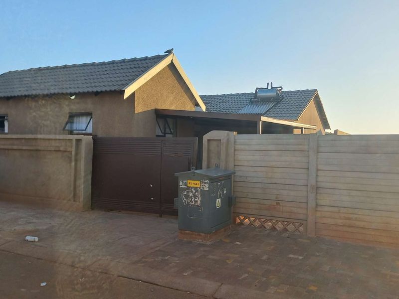 Front exterior view of a residential house featuring a tiled roof, solar water heater, and secure precast concrete boundary wall with a metal gate.