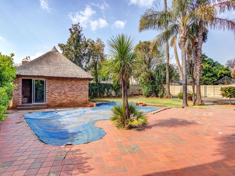 Outdoor pool area featuring a brick-paved patio, a thatched roof lapa, and a swimming pool with a cover, set within a private garden.