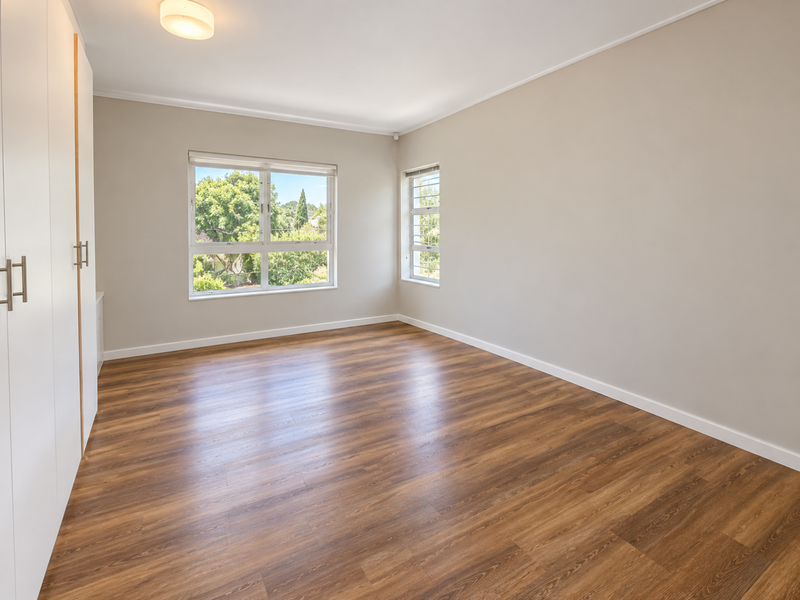 A bright, empty bedroom featuring modern laminate flooring, built-in cupboards, and large windows providing natural light and garden views.