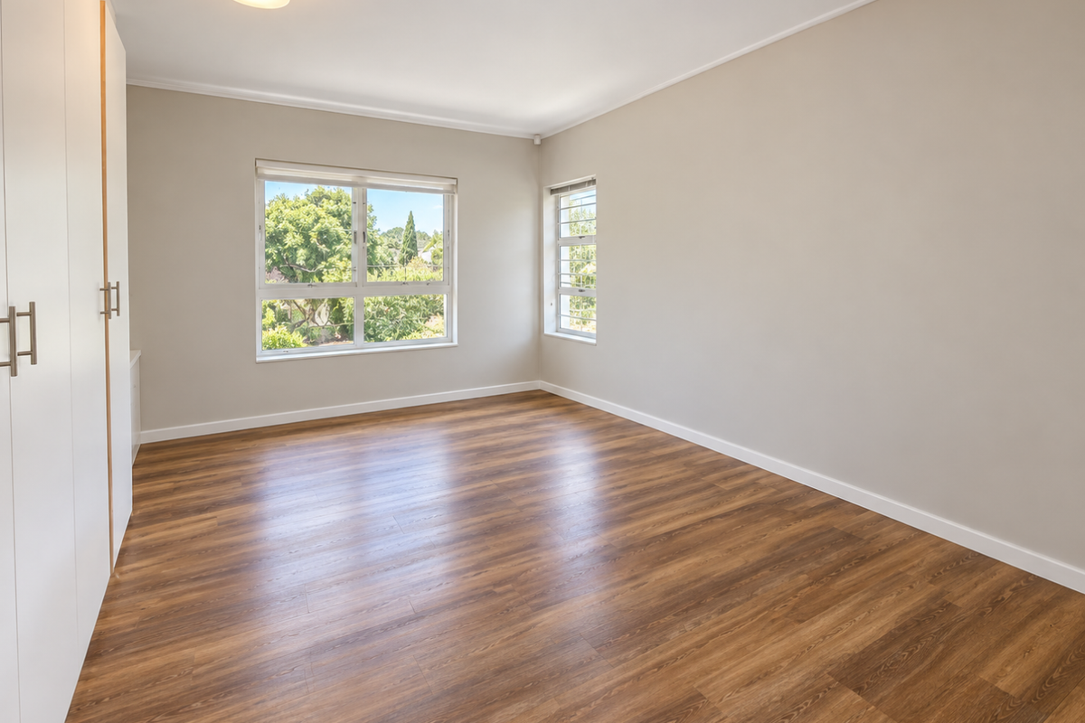 A bright, empty bedroom featuring modern laminate flooring, built-in cupboards, and large windows providing natural light and garden views.