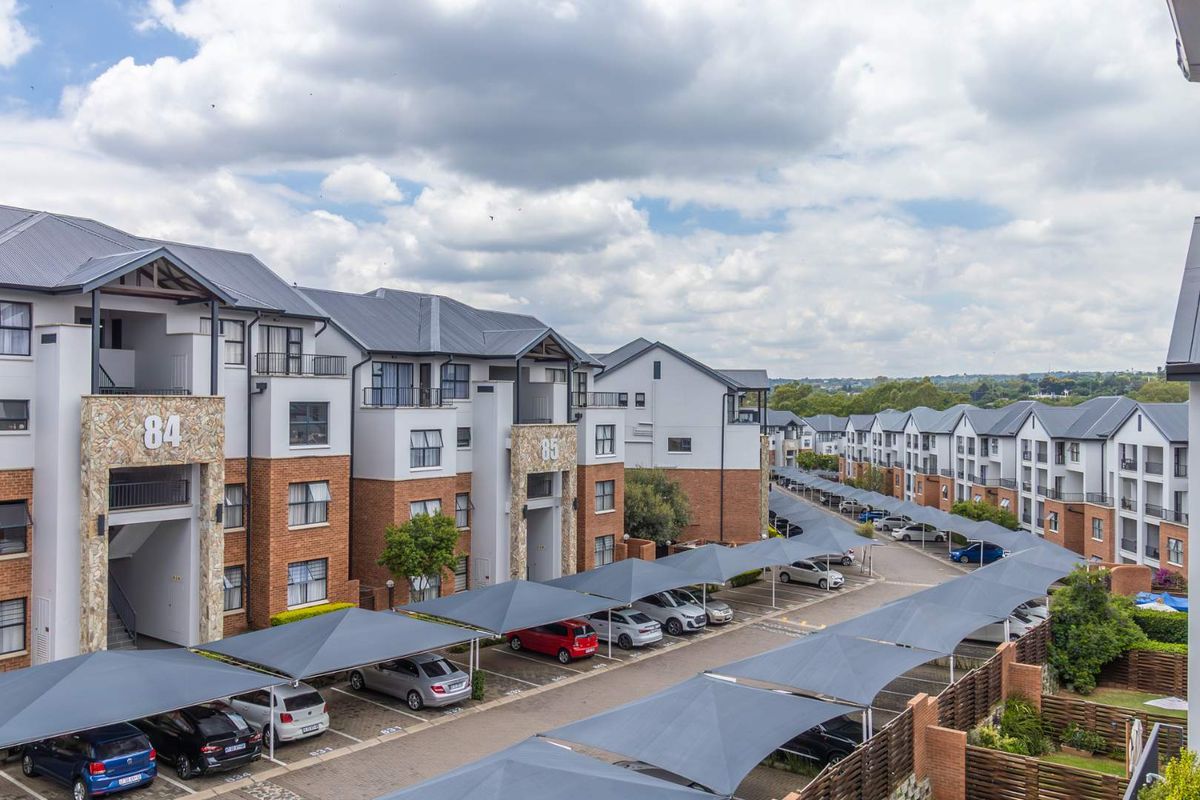 An elevated view of a modern apartment complex featuring multi-story residential buildings, paved parking areas with shade cloth carports, and landscaped surroundings.