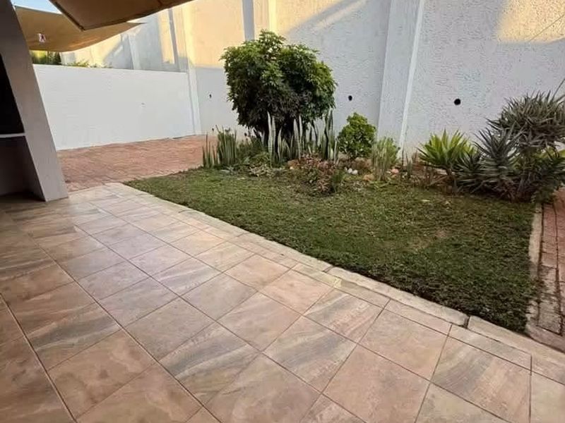 A covered patio area featuring tiled flooring, a retractable awning, and an adjacent landscaped garden bed against a high boundary wall.
