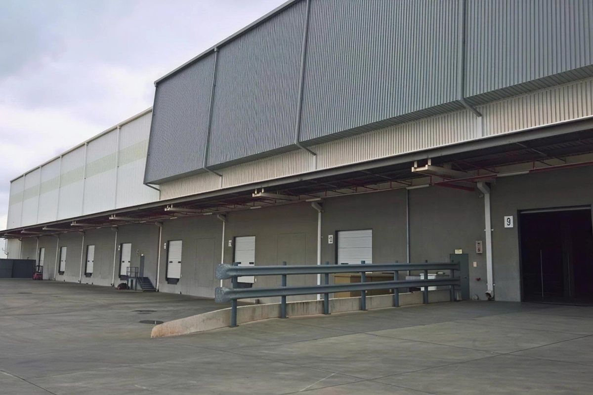 Industrial warehouse exterior featuring multiple loading docks with roller shutter doors, concrete hardstand area, and protective safety bollards under a covered canopy.