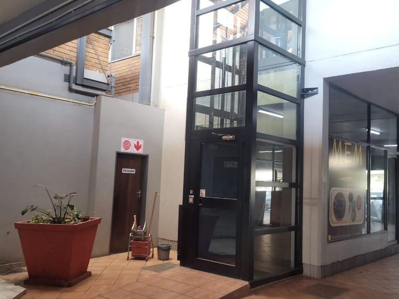Commercial building entrance area featuring a modern glass elevator shaft, tiled flooring, and access to storefronts.
