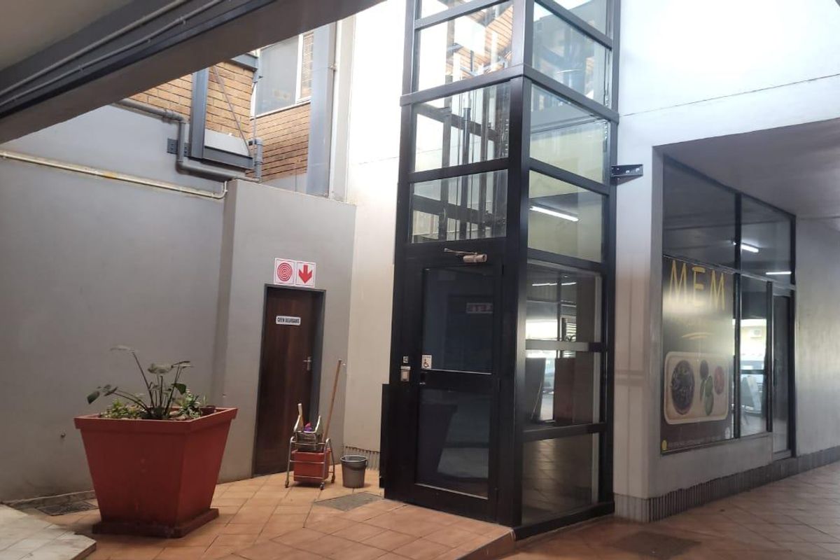 Commercial building entrance area featuring a modern glass elevator shaft, tiled flooring, and access to storefronts.