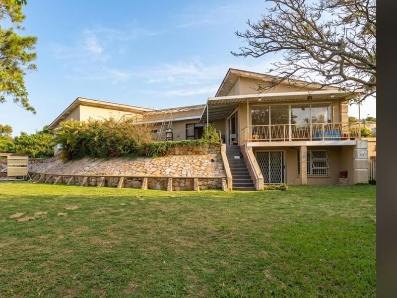 Rear view of the property featuring a stone retaining wall, elevated balcony with metal railing, and a spacious lawn area.