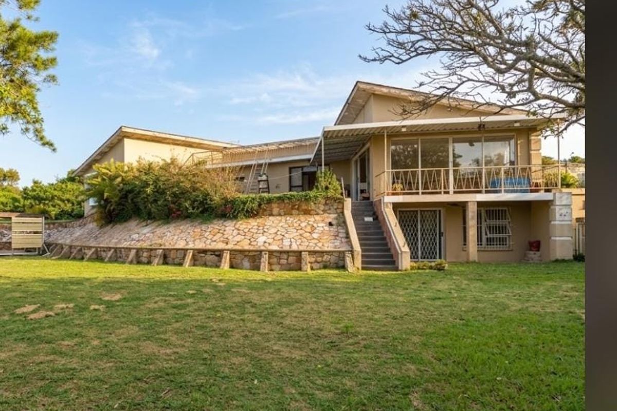 Rear view of the property featuring a stone retaining wall, elevated balcony with metal railing, and a spacious lawn area.