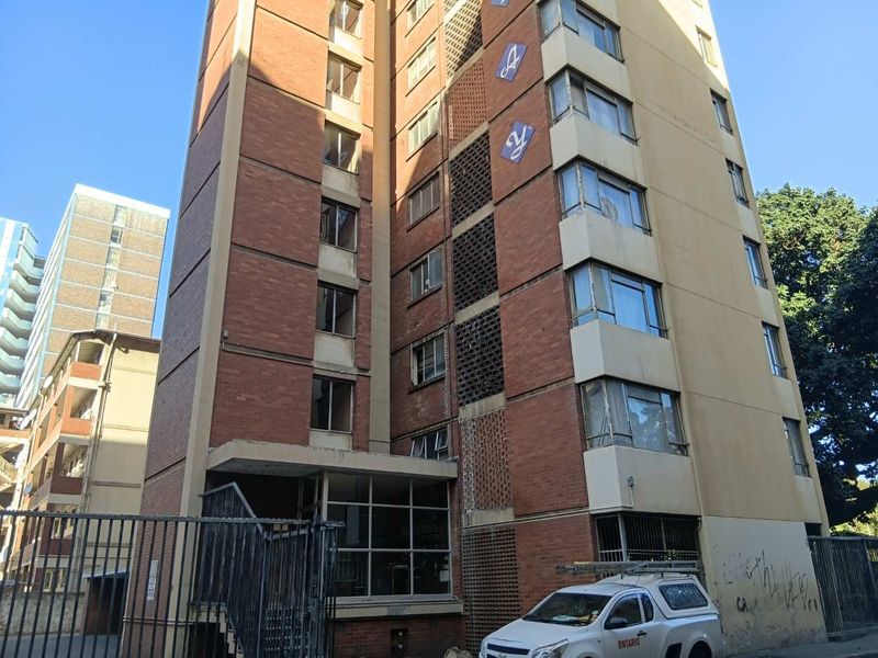 Exterior view of a multi-story apartment building featuring a brick facade, decorative breeze block detailing, and secure perimeter fencing.