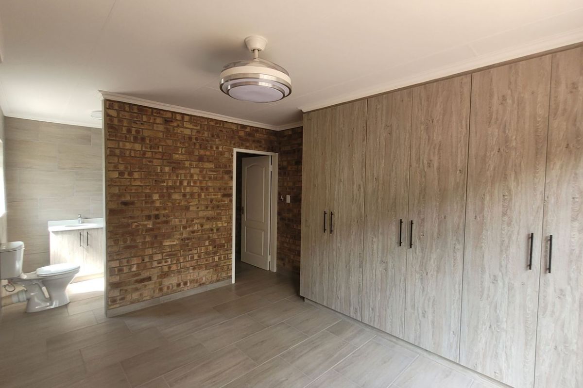 Spacious bedroom featuring built-in cupboards, an exposed brick feature wall, tiled flooring, and an integrated en-suite bathroom.