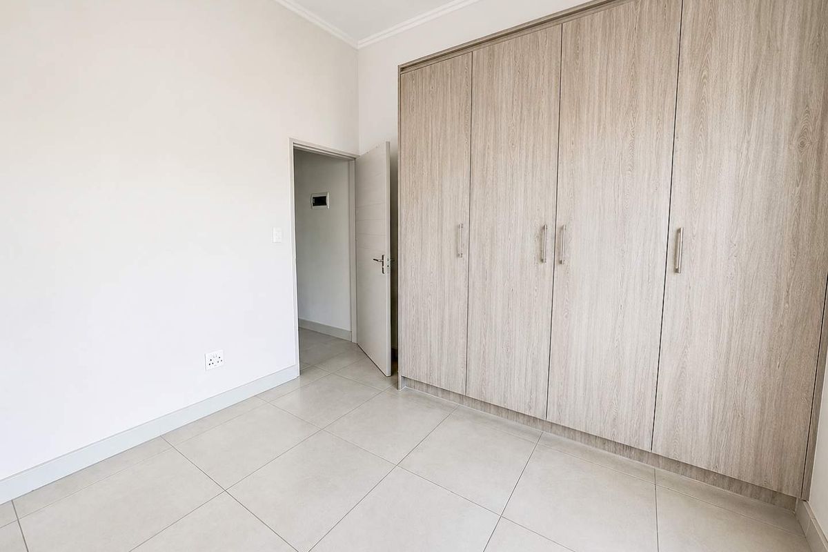 A bright, empty bedroom featuring modern light-toned tiled flooring and extensive built-in wooden cupboards, offering a clean and neutral living space.