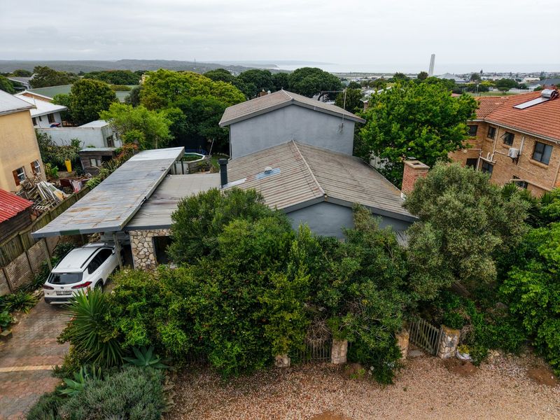 Aerial view of the property showing the main dwelling, corrugated metal roofing, attached carport, and surrounding garden area with coastal views in the distance.