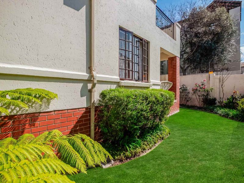 A well-maintained rear exterior view of the townhouse featuring a manicured lawn, brick and plaster facade, and a balcony with metal railings.