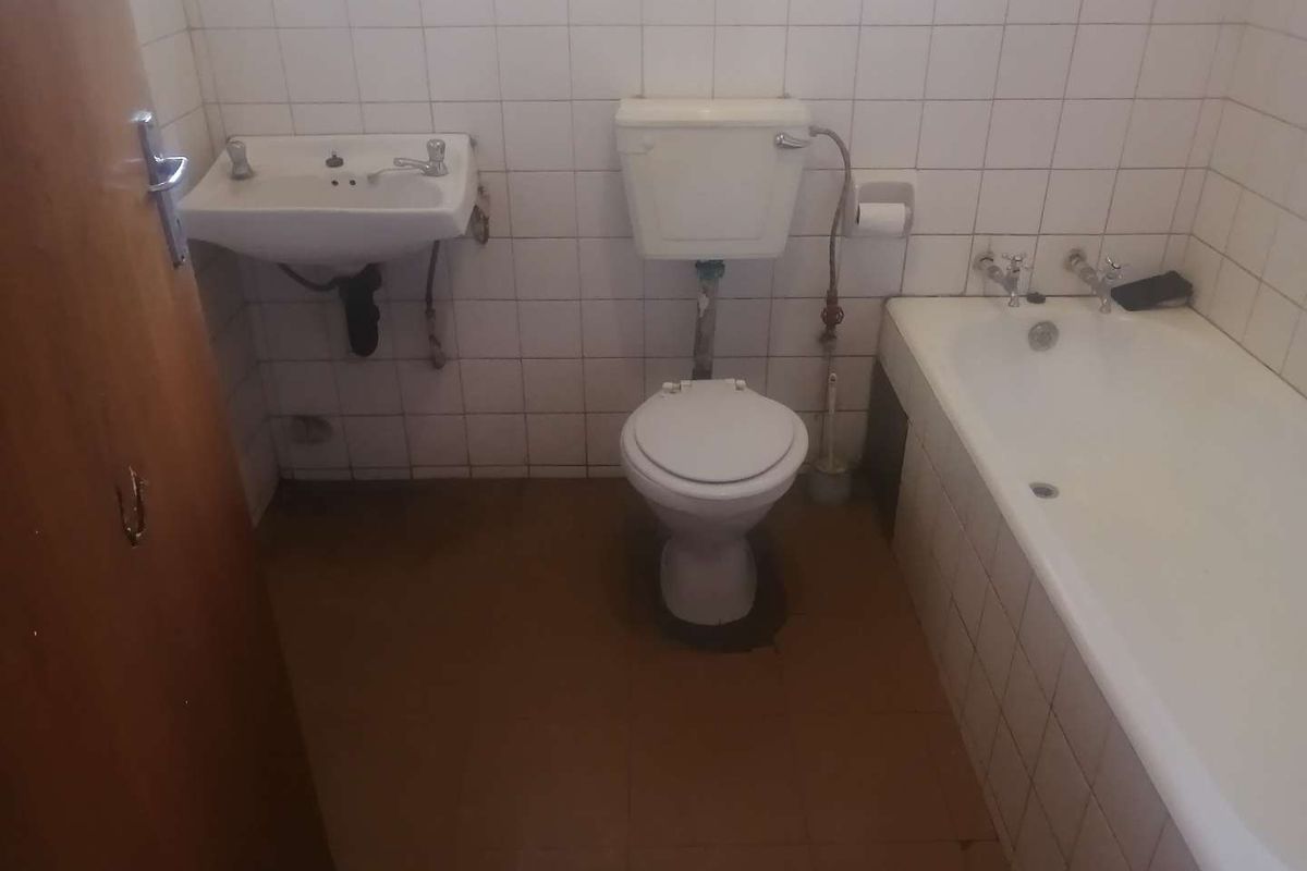 A dated bathroom featuring a standard bathtub, wall-mounted basin, and toilet with white wall tiles and brown floor tiles.