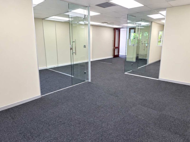 Modern office space featuring glass partition walls, carpeted flooring, and a suspended ceiling with recessed lighting, offering a clean and professional work environment.