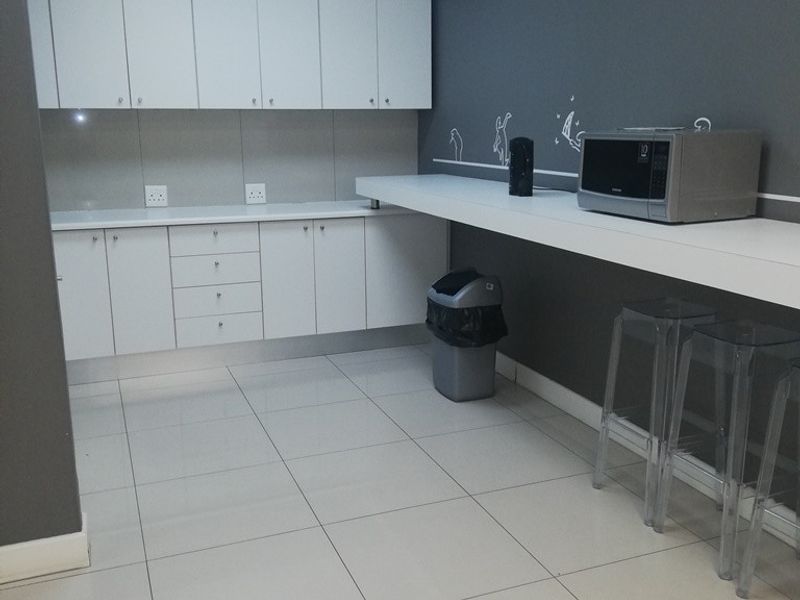 A functional office kitchenette featuring white built-in cabinetry, a long laminate countertop, and neutral ceramic floor tiles with recessed ceiling lighting.