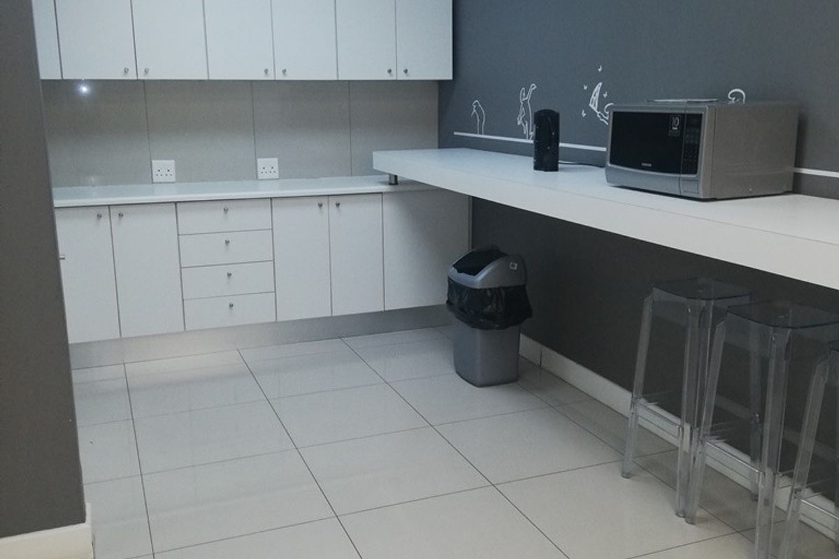A functional office kitchenette featuring white built-in cabinetry, a long laminate countertop, and neutral ceramic floor tiles with recessed ceiling lighting.