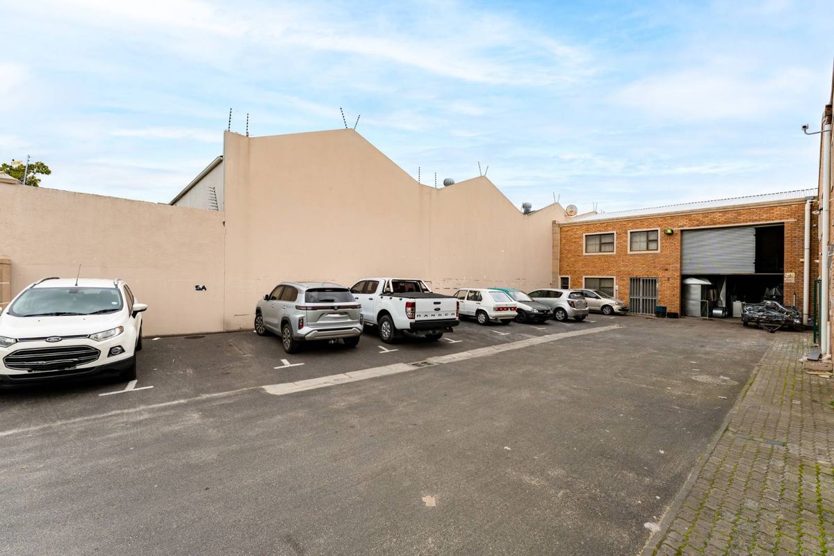 Exterior view of a mixed-use property featuring a paved parking area, perimeter boundary wall with security spikes, and a large industrial roller shutter door.
