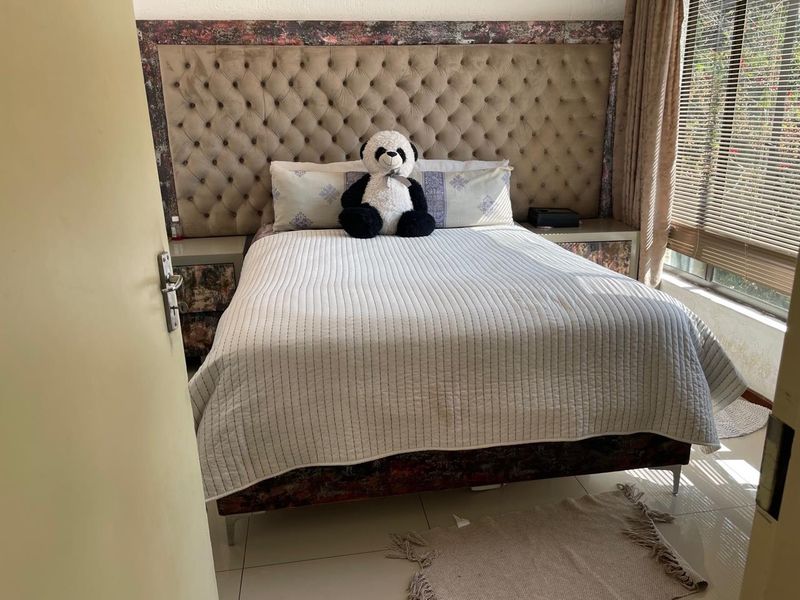 A bedroom featuring a large tufted headboard with integrated bedside tables, tiled flooring, and venetian blinds.