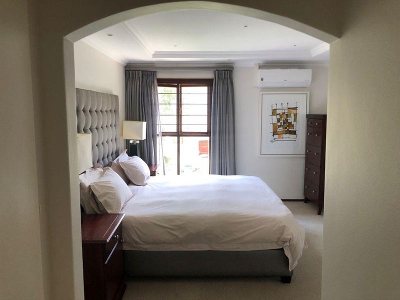 A well-maintained bedroom featuring recessed ceiling lighting, decorative cove molding, and a wall-mounted air conditioning unit, with access to natural light through a window.