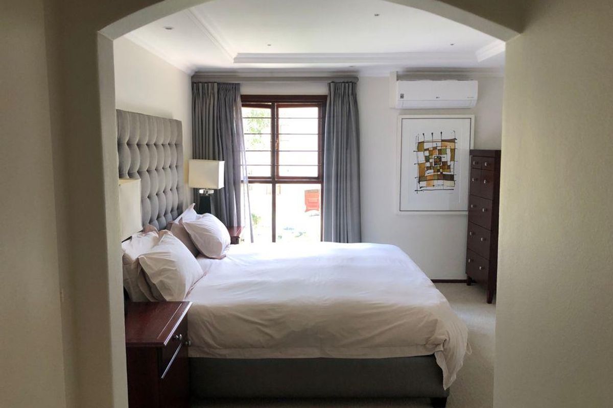 A well-maintained bedroom featuring recessed ceiling lighting, decorative cove molding, and a wall-mounted air conditioning unit, with access to natural light through a window.
