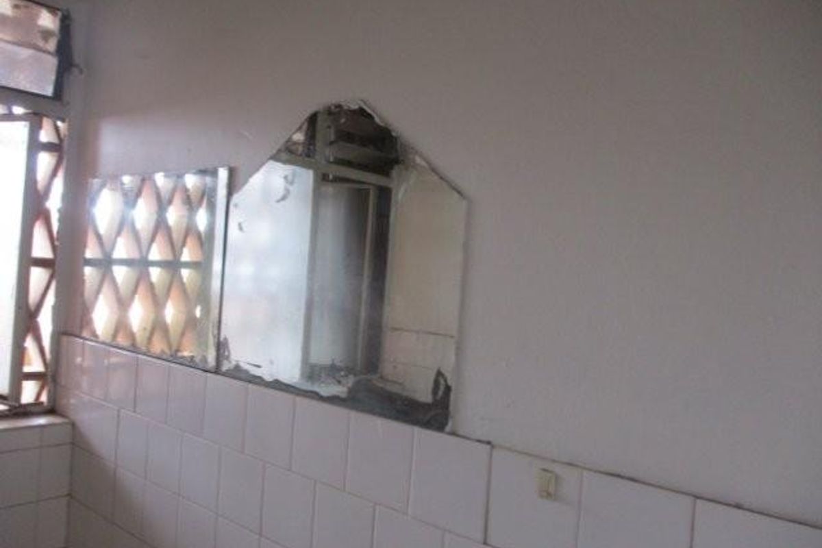A bathroom interior featuring wall tiles and a window with security bars, showing significant wear and damage to the wall-mounted mirror.