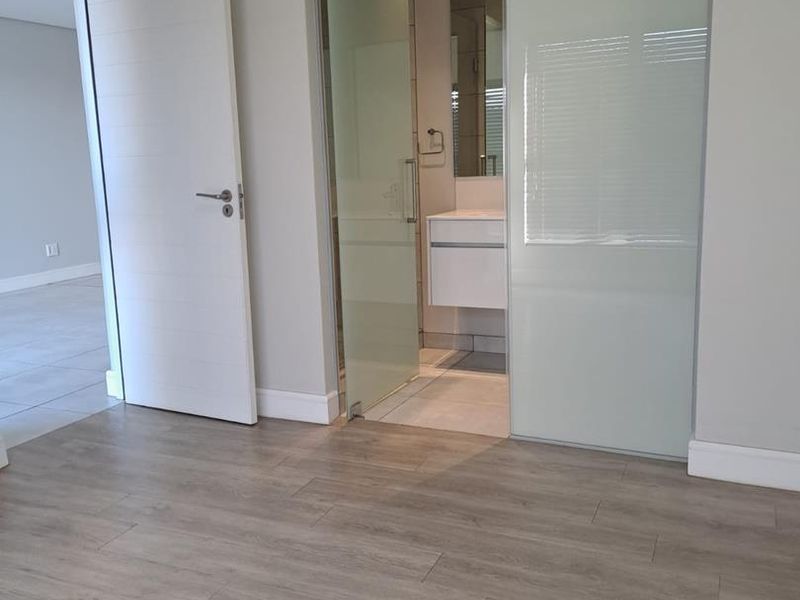 A bright, modern bedroom featuring light wood-look laminate flooring, neutral walls, and a sleek frosted glass sliding door leading to an en-suite bathroom.
