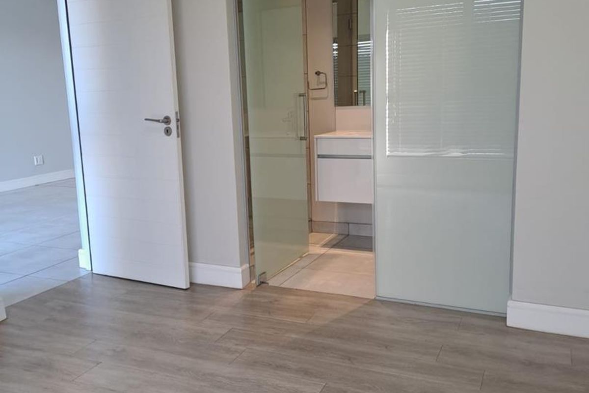 A bright, modern bedroom featuring light wood-look laminate flooring, neutral walls, and a sleek frosted glass sliding door leading to an en-suite bathroom.